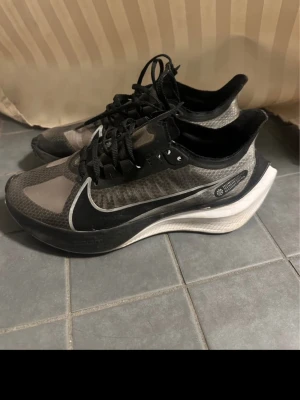 Nike ZoomX Vaporfly NEXT% 2 sneakers - Säljer ett par Nike ZoomX Vaporfly NEXT% 2 sneakers i svart och grått med vit sula. Skorna har en sportig design med genomskinligt meshmaterial, stora vita swoosh-loggor och avancerad dämpning i sulan. Perfekta för dig som gillar löpning och streetstyle. Använda 1 gång. 