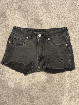Svarta jeansshorts med kant - Säljer ett par svarta jeansshorts med fransig kant och slitningar framtill. Klassisk femficksmodell med knapp och dragkedja. Perfekta för en avslappnad stil.