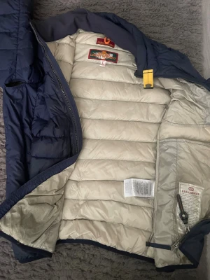 Mörkblå dunjacka från Parajumpers - Snygg mörkblå dunjacka från Parajumpers med quiltad design och gul detalj vid halsen. Jackan har två dragkedjeförsedda fickor framtill och patch på ärmen. Insidan är ljus och jackan har hög krage. Perfekt för kalla vinterdagar.