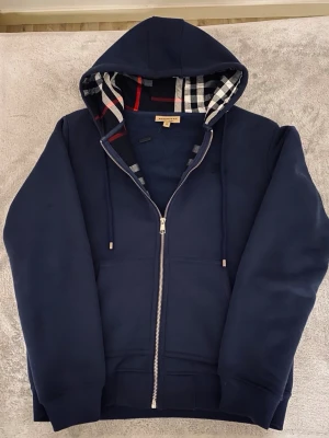 Marinblå hoodie från Burberry - Snygg marinblå hoodie från Burberry med dragkedja och klassiskt rutigt foder i huvan. Hoodien har dragsko i huvan, ribbade muddar och två fickor framtill. Perfekt för dig som gillar stilrena och exklusiva plagg med en twist.