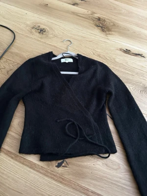 Svart omlottkofta från Gina Tricot - Säljer en svart stickad omlottkofta från Gina Tricot med långa ärmar och knytband i midjan. Koftan har en mjuk och lite luddig yta, perfekt för lager på lager och en snygg vardagslook.