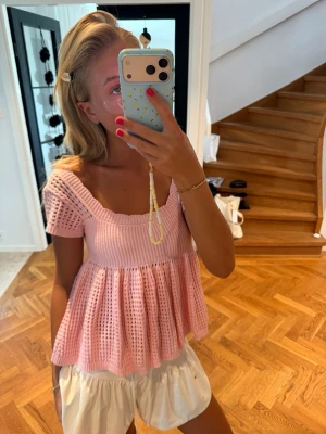 Ljusrosa stickad topp med volang - Supersöt ljusrosa stickad topp med korta ärmar och volang nedtill. Toppen har en fyrkantig ringning och ett luftigt, hålmönstrat material som ger en somrig vibe. Perfekt att styla med shorts eller kjol för en chill look.
