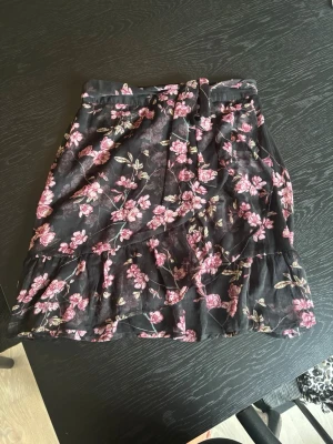 Svart blommig kortkjol med volang - Svart blommig kortkjol med rosa blommor och volangdetaljer. Kjolen har omlottkänsla och ett knytband i midjan. 