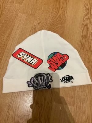 SYNA WORLD skull cap/mössa - (rep) Vit Syna world skull cap, fint skick men har en pytte liten fläck, går nog att ta bort i handtvätt men ej försökt själv. kommer med stickers och påsen om jag hittar