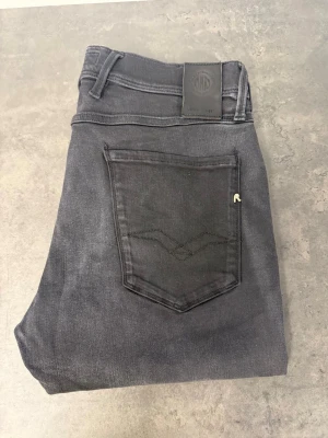 Svarta slim fit jeans Replay Anbass 33/34 - Snygga svarta slim fit jeans från Replay med klassisk femficksdesign och diskret logga på bakfickan. Jeansen har en smal passform och är tillverkade i stretchigt denimtyg för extra komfort. Perfekta för en trendig och avslappnad look.