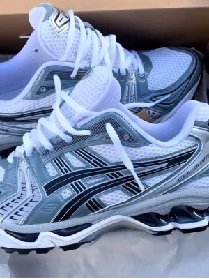 Asics vita och silver sneakers - Säljer ett par fräscha Asics sneakers i vitt och silver med svarta detaljer. Skorna har mesh-ovandel för bra ventilation, klassisk Asics-logga på sidan och tjock sula för skön komfort. Perfekta för dig som gillar sportig och trendig stil.
