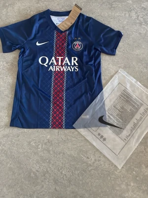 PSG matchtröja blå Nike - Snygg blå PSG matchtröja från Nike med Qatar Airways-tryck på bröstet och klubbmärke. Tröjan har korta ärmar, Dri-FIT-material och ett rött och vitt mönster längs mitten. Perfekt för fotbollsträning eller att bära som supporter.