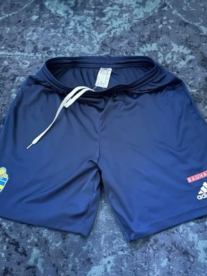 Blåa SvFF shorts från Adidas - Mörkblå sportshorts från Adidas med Svenska Fotbollförbundets märke och Bauhaus-logga. Tillverkade i lätt och snabbtorkande material, har dragsko i midjan för bästa passform. Perfekta för fotbollsträning eller match. 