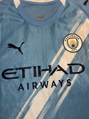 Manchester City matchtröja Haaland #9 - Snygg ljusblå Manchester City matchtröja från Puma med Haaland och nummer 9 på ryggen. Tröjan har klubbmärke, sponsorlogga och svarta detaljer. Perfekt för fotbollsfans som vill stötta sitt lag på match eller träning.