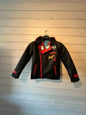 Spider-Man  jacka med huva - Barn  - Cool svart Spider-Man cosplay jacka med röda detaljer och tryck. Jackan har en röd huva med spindelnätsmönster och vita ögon, samt tryck fram och bak med text och spindelmotiv. Perfekt för maskerad eller cosplay. 7-8 år. Strl 128 . 
