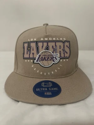 Beige Lakers keps Ultra Game NBA - Snygg beige keps med rak skärm från Ultra Game, prydd med stort Los Angeles Lakers-tryck i lila och vitt framtill. Klassisk snapback-modell med broderad logga på sidan och officiell NBA-märkning. Perfekt för dig som älskar basket och streetwear.