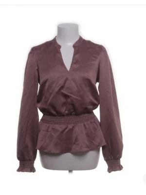 Blus - Snygg plommonlila blus i glansigt material med smock i midjan och volang nedtill. Blusen har v-ringning och långa ärmar med smockade muddar. Perfekt för dig som vill ha en trendig och feminin look.