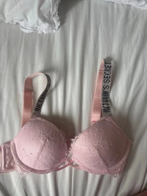Rosa spets-bh från Victoria's Secret - Söt ljusrosa bh från Victoria's Secret med vadderade kupor och spetsdetaljer. Axelbanden är dekorerade med glittrande text och ger en exklusiv känsla. Bh:n har bygel och justerbara axelband för bästa passform.