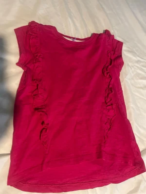 Rosa topp med volang från Sweet Me barn 122/128 - Supersöt rosa topp från Sweet Me med volangdetaljer längs sidorna och en liten rosett i halsen. Toppen är ärmlös och har en mjuk, bekväm känsla som passar perfekt till jeans eller kjol. Barn storlek 122/128