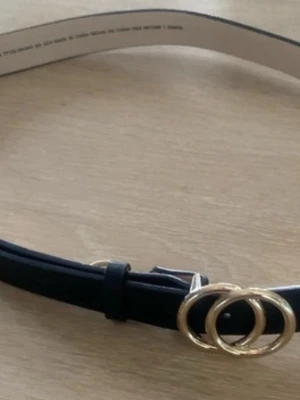 Svart bälte med guldfärgat spänne - Snyggt svart bälte med ett stilrent guldfärgat dubbelring-spänne. Perfekt accessoar för att lyfta vilken outfit som helst. Bältet har en klassisk design och är gjort i ett slätt material.