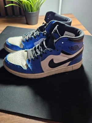 Nike Air Jordan 1 High blå/vit - Säljer ett par Nike Air Jordan 1 High sneakers i blått, vitt och svart. Klassisk high-top siluett med svart swoosh, svarta skosnören och detaljerad vinge-logga på sidan. Ovandelen är i läder och har perforerad tåbox för extra ventilation.