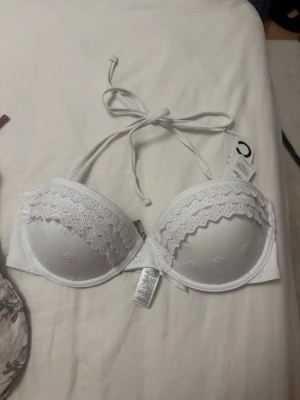 Vit bikini med broderade detaljer - Söt vit bikini med vadderade kupor och broderade spetsdetaljer i blommönster. Kuporna har vågiga kanter och knytning i nacken för en feminin look. Det går även att ta bort banden och ha den som en bandeau bikini. Perfekt för dig som gillar romantiska underkläder. Den är aldrig använd då den inte passar mig, men den är verkligen jättefin. Storleken är 38 men skulle helt klart säga 75C.