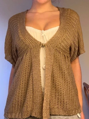 Beige stickad kortärmad kofta - Säljer en beige stickad kofta med korta ärmar och öppen front. Koftan har en lös och luftig passform med bred ribbstickad kant runt halsen och framtill. Perfekt att slänga över ett linne för en avslappnad look.