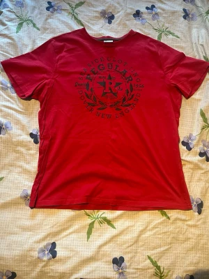 Röd t-shirt med tryck från Regular - Snygg röd t-shirt från Regular med svart tryck framtill som har text och stjärndetaljer. Klassisk rund halsringning och korta ärmar. Perfekt för en avslappnad stil och enkel att matcha med jeans eller shorts.