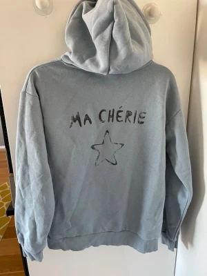 Ljusblå hoodie med text och stjärna - Ljusblå hoodie med huva och trycket 'MA CHÉRIE' samt en stjärna både fram och bak. Mjuk och skön, perfekt för chill dagar. Ribbstickade muddar vid ärmslut och nederkant. Använd 1 gång 
