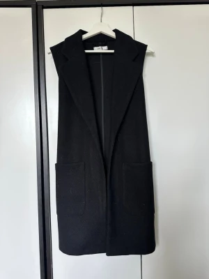 Svart lång väst med fickor och knytband - Stilren svart lång väst med breda slag, två stora framfickor och knytband i ryggen. Västen är ärmlös och har en rak passform som ger en clean look. Perfekt att slänga över en hoodie eller skjorta för en extra touch. Materialet ser ut att vara tjockt och mjukt, troligen ullblandning.