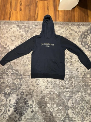 Mörkblå hoodie från Jack & Jones - Snygg mörkblå hoodie från Jack & Jones med tryck 'Jack & Jones c1990' på bröstet. Klassisk huva och stor magficka framtill. Tillverkad i mjukt material som är skönt att ha på sig. Perfekt för en avslappnad stil.