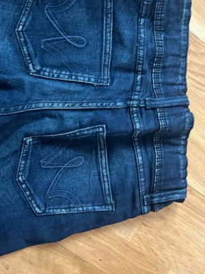 Mörkblå stretchjeans med broderade fickor och justerbar i midjan  - Snygga mörkblå jeans med stretch och dubbla knappar i midjan. Bakfickorna har coola broderade detaljer och passformen är slim. Perfekta för dig som gillar en klassisk men ändå trendig look.