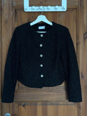 Svart cardiganjacka från Vila - Stilren svart cardiganjacka från Vila med glittrande struktur och dekorativa pärlknappar i blomform. Jackan har rund halsringning, lång ärm och en något kortare, rak passform. Perfekt för att lyfta din outfit med en elegant touch.