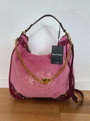 Juicy Couture rosa axelremsväska - Juicy Couture handväska i rosa sammet med mörkrosa detaljer och guldfärgade kedjor. Väskan har ett hjärtformat hängsmycke och ett matchande litet fodral med Juicy Couture-logga. Snygg och rymlig modell med både handtag och axelrem.