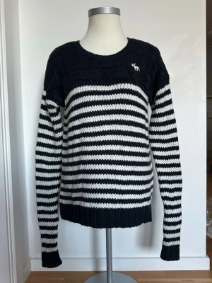 Randig stickad tröja från Abercrombie - Snygg stickad tröja från Abercrombie & Fitch i svart och vitt med horisontella ränder. Tröjan har rund hals, långa ärmar och en liten broderad älg-logga på bröstet. Perfekt för dig som gillar klassisk och avslappnad stil. Kan vara nopprig på vissa ställen.