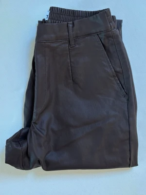 Bruna fuskskinnbyxor med resår - Snygga bruna byxor i fuskskinn med resår i midjan och bälteshällor. Byxorna har raka ben och sidofickor för en clean look. Perfekta för dig som vill ha en trendig och bekväm stil. Aldrig använda men lappen är av 💖