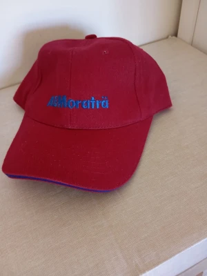 Röd keps med blå brodyr - Säljer en röd keps från AB Moraträ med böjd skärm och blå broderad logga framtill. Klassisk modell med justerbar rem bak. Tillverkad i bomullstyg som känns både slitstarkt och bekvämt. Perfekt för dig som vill ha en enkel men snygg accessoar.