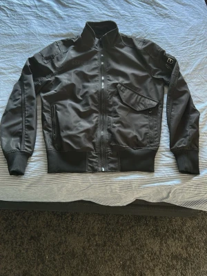 Svart bomberjacka från G-Star RAW - Snygg svart bomberjacka från G-Star RAW med dragkedja framtill, ribbade muddar och hög krage. Jackan har en sned bröstficka med lock, sidoficka med dragkedja och patch med logga på ärmen. Materialet är glansigt och ger en modern look.