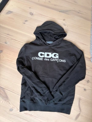Svart CDG hoodie Comme des Garçons - Snygg svart hoodie från Comme des Garçons med stor vit CDG-logga och texten 'COMME des GARÇONS' på bröstet. Klassisk känguruficka framtill och huva. Perfekt för dig som gillar streetwear och vill ha en stilren look.