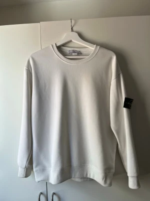 Vit sweatshirt från Stone Island - Snygg vit sweatshirt från Stone Island med klassisk rund hals och ribbade muddar. Tröjan har den ikoniska svarta patchen med gult och grönt Stone Island-märke på vänster ärm. Perfekt för en clean och stilren look.