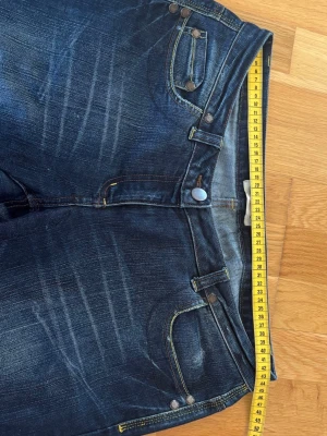 Mörkblå lågmidjade bootcut jeans med slitningar - Säljer ett par mörkblå bootcut jeans med snygga slitningar och kontrastsömmar. Klassisk femficksmodell med knappgylf och nitar. Jeansen har en rak passform över låren och lätt utsvängda ben. Perfekta för en avslappnad och trendig look.