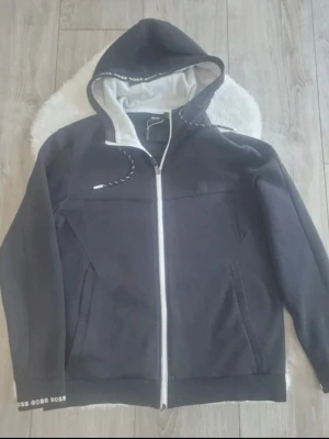 Svart hoodie från Hugo Boss med dragkedja - Snygg svart hoodie från Hugo Boss med vit dragkedja och logga på bröstet. Huvan har snörning och insidan är ljusgrå. Ärmslutet har diskret BOSS-tryck. Perfekt för en avslappnad och stilren look.