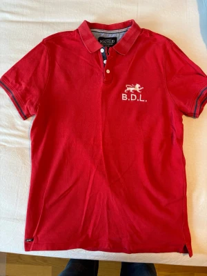 Röd pikétröja från Bonde Ltd - Röd pikétröja från Bonde Ltd med vit broderad logga och texten B.D.L. på bröstet. Tröjan har klassisk krage, knappar framtill och korta ärmar med randiga detaljer vid ärmsluten. Tillverkad i bomull för en bekväm känsla.