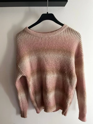 Pastellrosa stickad tröja - Mysig stickad tröja i pastellrosa och beige toner med rund hals och avslappnad passform. Tröjan har ett unikt batikmönster och ribbade muddar vid ärmslut och nederkant. Perfekt för dig som gillar en chill och trendig stil.