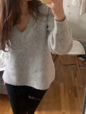 Ljusgrå stickad tröja från AWARE - Mysig ljusgrå stickad tröja från AWARE med v-ringning och långa ärmar. Tröjan har en mjuk, ribbstickad struktur och avslappnad passform som passar perfekt till jeans eller leggings.