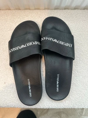 Svarta Emporio Armani sandaler - Snygga svarta sandaler från Emporio Armani med vitt logotyptryck över remmen. Tillverkade i syntetmaterial med en klassisk slip-in design och räfflad sula för extra grepp. Perfekta för en avslappnad och trendig look.