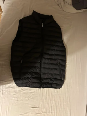 Svart dunväst med dragkedja - Snygg svart dunväst med horisontella sömmar och hög krage. Västen har två fickor med dragkedja framtill och stängs med en hel dragkedja. Perfekt för lager-på-lager och enkel att matcha till olika outfits.