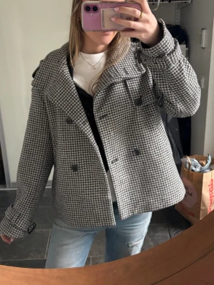 Rutig skepparkavaj i ullmix - Snygg rutig skepparkavaj i grått och vitt med dubbelknäppning och stora svarta knappar. Jackan har lång ärm, axelklaffar och klassisk krage. Materialet känns som en ullmix och passformen är loose för en chill vibe.