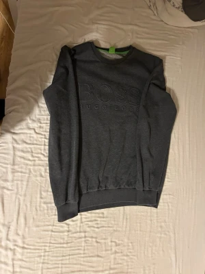 Mörkgrå sweatshirt från Hugo Boss - Är storlek xl men sitter som L-M Mörkgrå sweatshirt från Hugo Boss med diskret broderad logga på bröstet. Tröjan har rund halsringning, ribbade muddar vid ärmslut och nederkant samt en klassisk, avslappnad passform. Perfekt för en stilren och enkel look.
