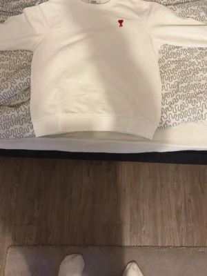 Vit sweatshirt från Ami Paris - Står XL men S , Säljer en stilren vit sweatshirt från Ami Paris med det klassiska röda hjärtemblemet broderat på bröstet. Tröjan har rund halsringning, långa ärmar och ribbade muddar. Perfekt för en clean och trendig look. 