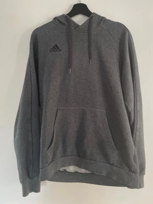 Adidas hoodie – M | grå | skönt material - Grå hoodie från Adidas i storlek M. Mjukt och skönt material med fleece-insida – perfekt för både vardag och träning   Klassisk modell med luva, snören och magficka samt Adidas-logga på bröstet.  Använd men fortfarande i bra skick. Finns lite mindre tecken på användning (se bilder), inget som påverkar funktionen.