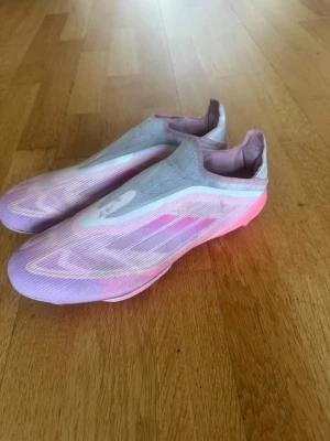 Adidas rosa/lila fotbollsskor utan snörning - Säljer ett par Adidas fotbollsskor i snygg rosa och lila färg med vita detaljer. Skorna har en strömlinjeformad design utan snörning och är tillverkade i ett lätt syntetmaterial för bästa känsla på planen. Perfekta för dig som vill sticka ut på matchen!