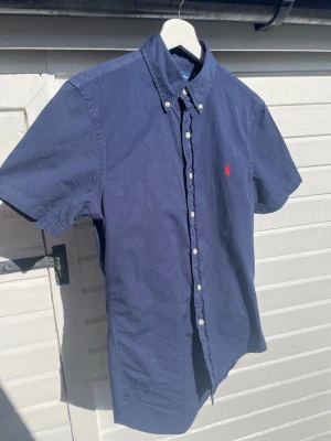 Marinblå kortärmad skjorta Polo Ralph Lauren - Snygg marinblå skjorta från Polo Ralph Lauren med korta ärmar och klassisk button-down krage. Skjortan har vita knappar framtill och den ikoniska röda logotypen broderad på bröstet. Tillverkad i mjuk bomull, perfekt för en clean och stilren look.