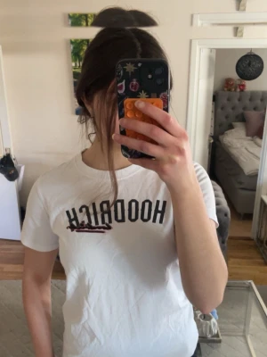 Vit HOODRICH t-shirt med tryck - Säljer en vit t-shirt från HOODRICH med svart tryck framtill och en röd detalj över loggan. Den är stor i storlek ,T-shirten har rund halsringning och korta ärmar. Perfekt för dig som gillar streetwear och vill ha en enkel men snygg look.