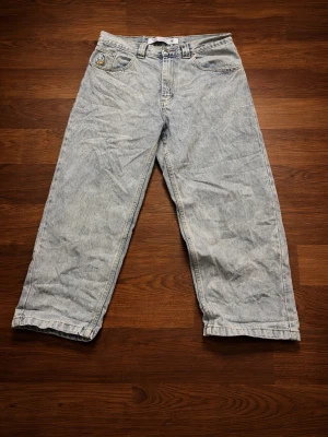 Ljusblå baggy jeans från OJ - Säljer ett par ljusblå baggy jeans från OJ med raka ben och klassisk femficksdesign. Jeansen har en gul logga på bakfickan och är tillverkade i denim. Perfekta för en avslappnad och trendig stil.